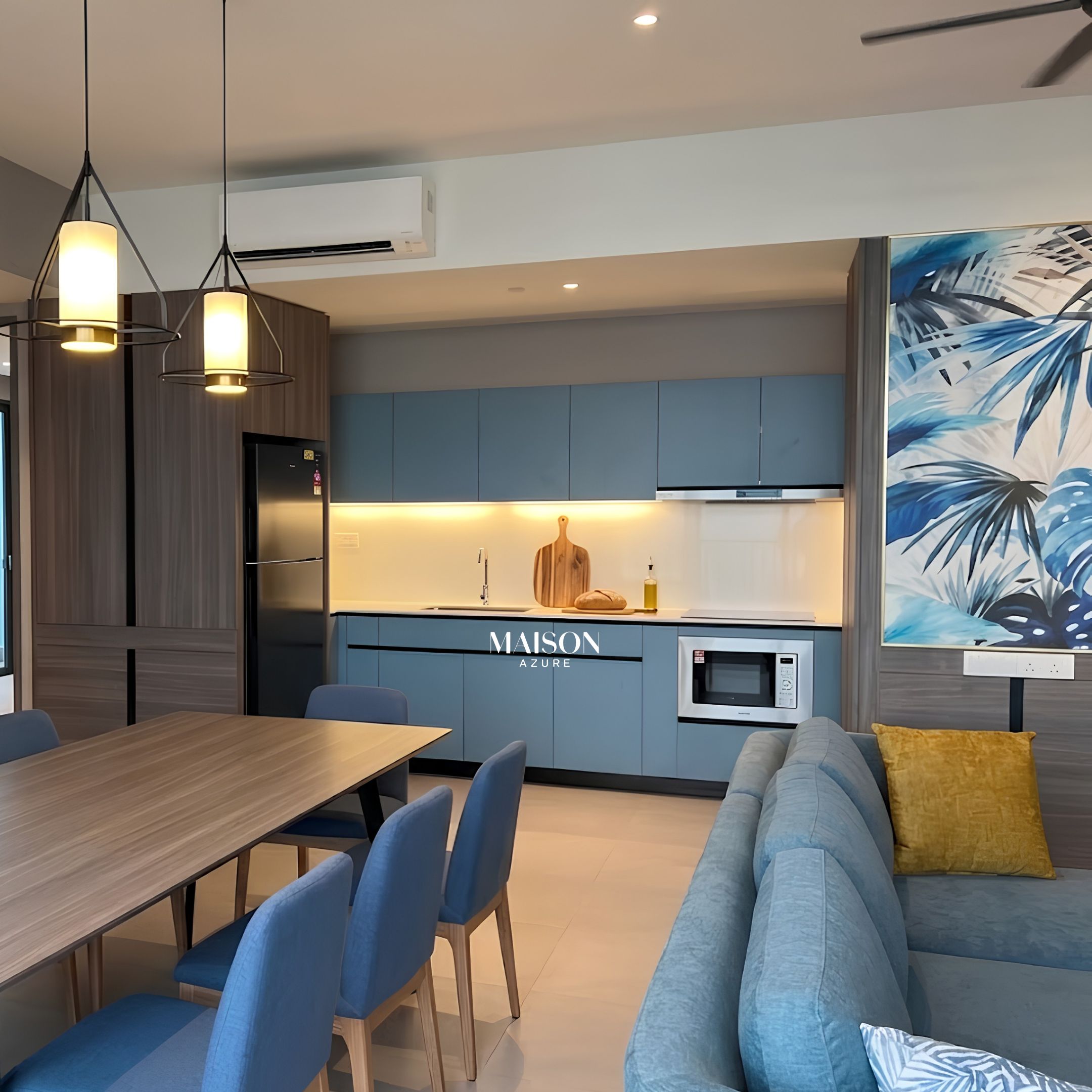 Une cuisine moderne avec des meubles bleus, un coin repas avec une table en bois et des chaises bleues, et un canapé gris agrémenté d’un coussin jaune.