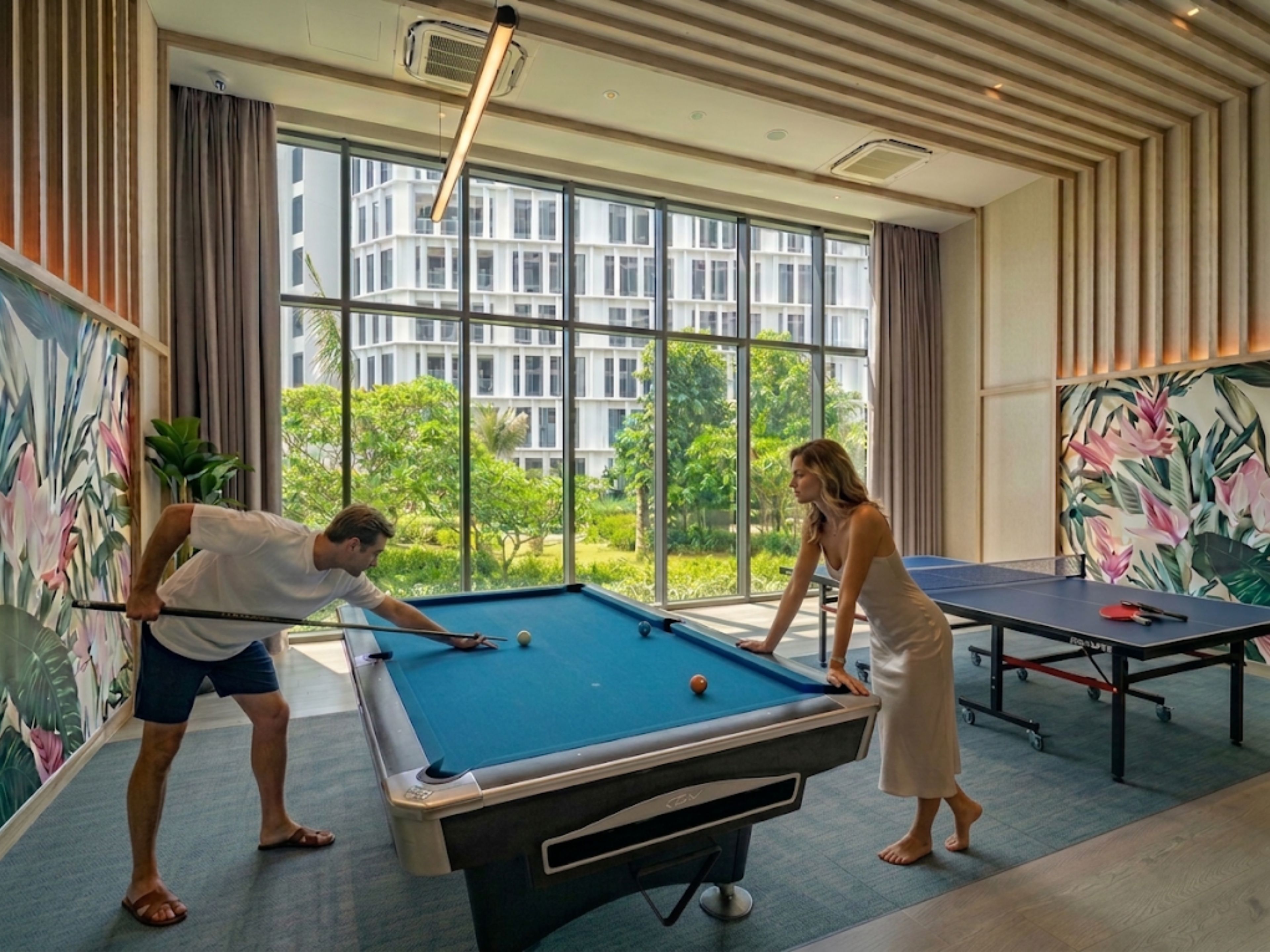 Una mesa de billar en una habitación con ventanas amplias, vistas al verdor exterior y un mural en la pared.