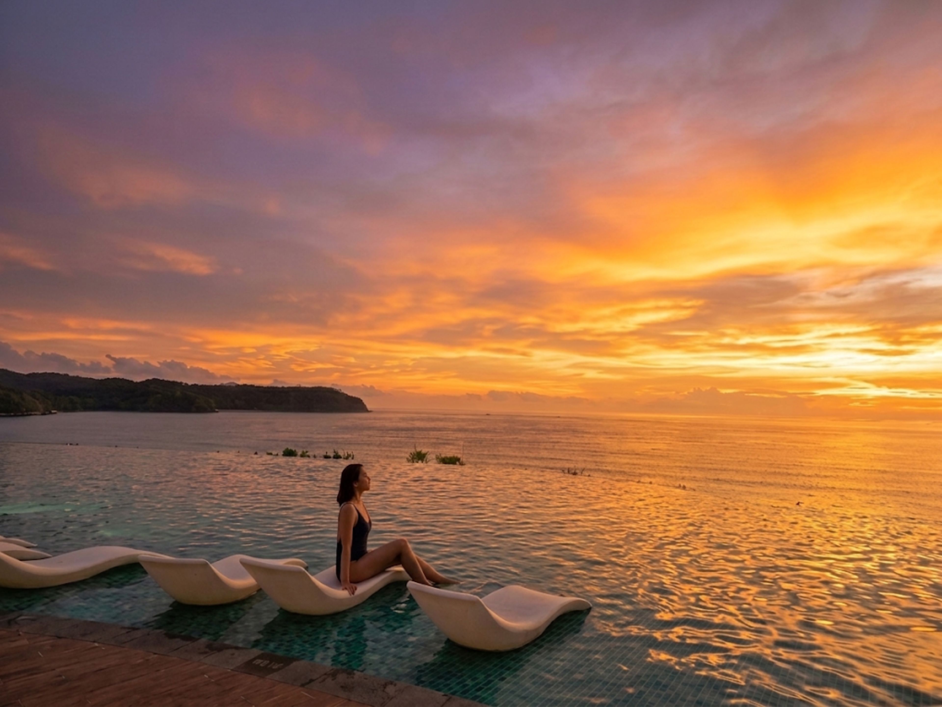 A person sitting on a floating lounge chair by a pool during sunset.