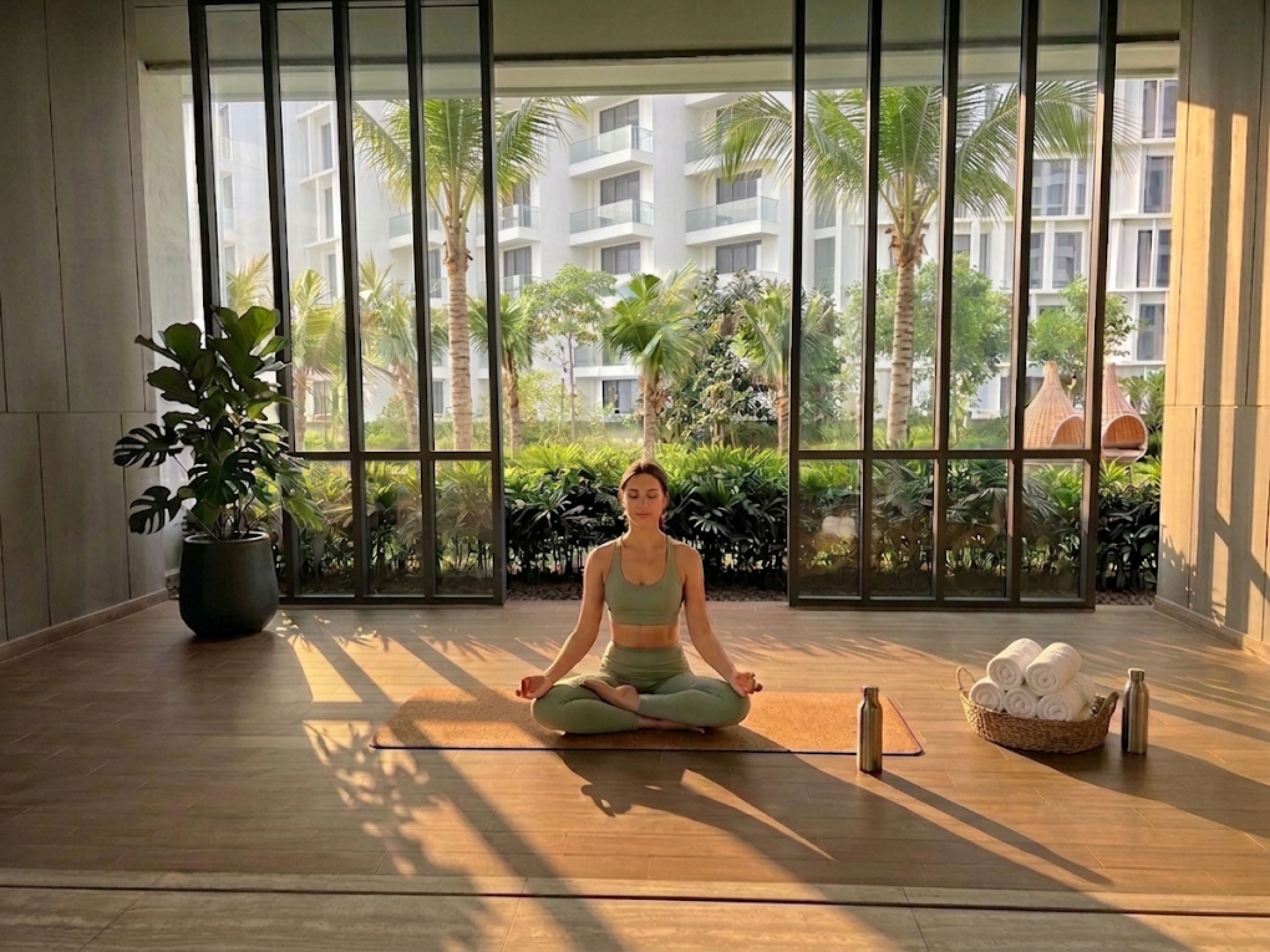 A person seated in a meditative pose on a mat inside a room with large windows.