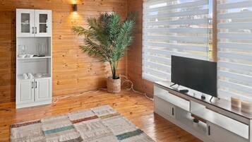 A room with wooden walls, a white cabinet with glass doors, a potted plant, a television on a wooden stand, and a window with blinds.