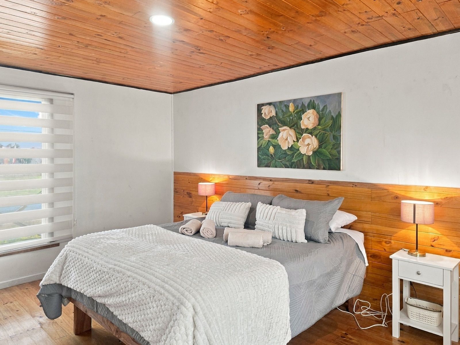 Een slaapkamer met een houten plafond, een bed met een grijze spreidek en witte kussens, een schilderij van bloemen aan de muur en een nachtkastje met een lamp.