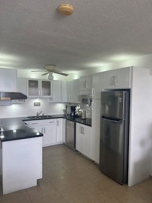 A modern kitchen with white cabinets, stainless steel appliances, and a ceiling fan. - Sunset Beach Unit 10 St Lawrence Gap Condos (Oistins)
