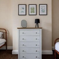Un dormitorio con cama, cómoda y tres láminas enmarcadas con motivos botánicos en la pared.