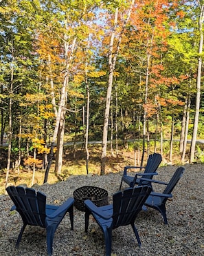 A forested area with a seating arrangement of chairs and a fire pit. - Cozy Cove! In-town & Convenient! (Ellsworth)