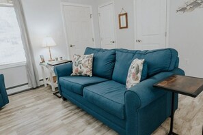 Un sofá color turquesa con cojines estampados, una mesita auxiliar y una lámpara en una habitación con puertas blancas y suelo de madera.