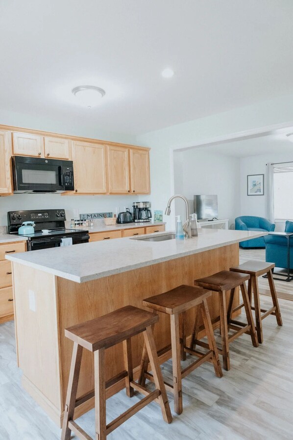 A modern kitchen with a central island, wooden stools, and a built-in microwave. - Cozy Cove! In-town & Convenient! (Ellsworth)