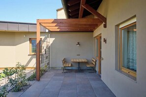 Ein überdachter Außenbereich mit Tisch und Bänken, einer Holz-Pergola und einem Gebäude mit großen Fenstern.
