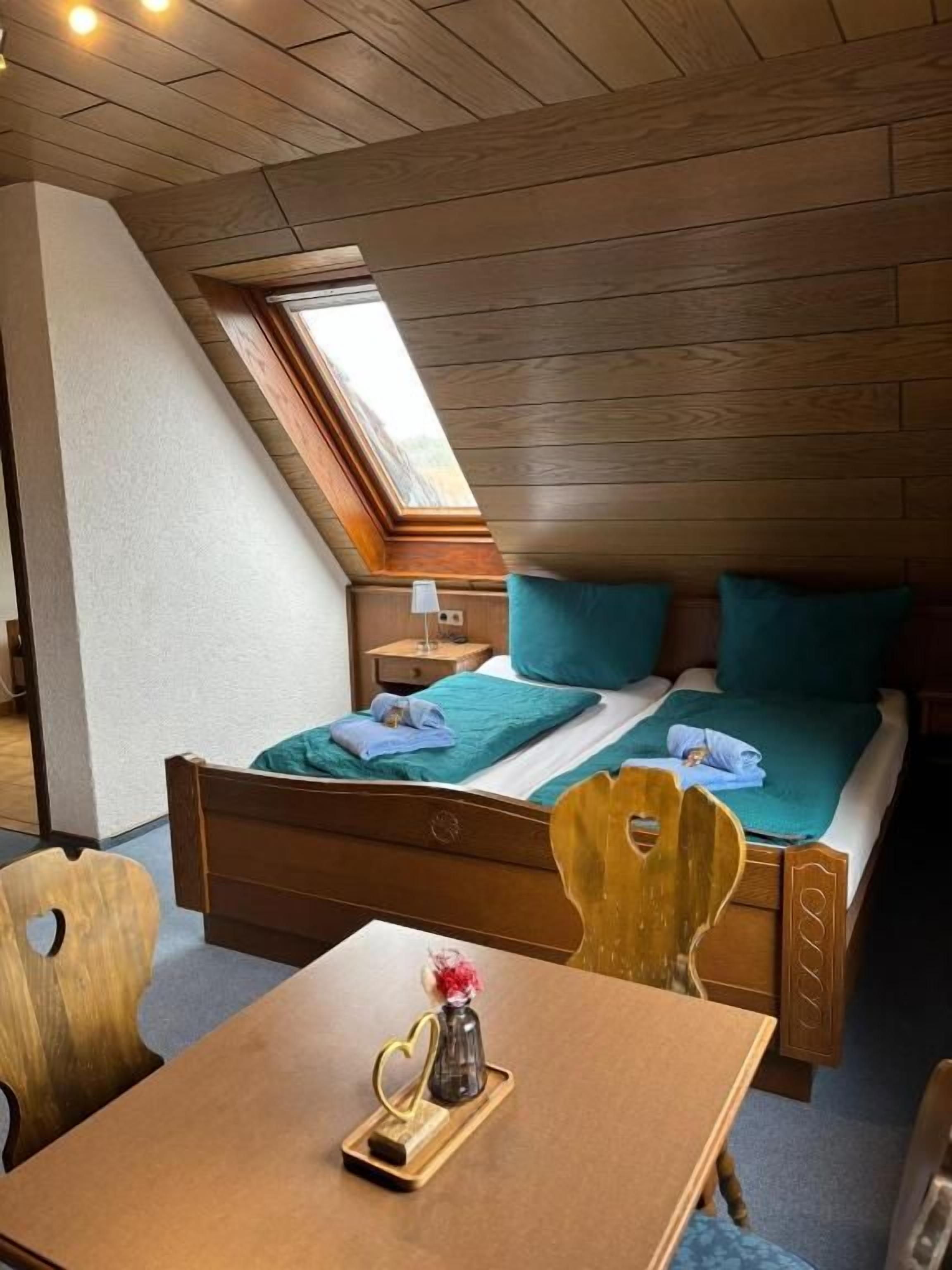 Een slaapkamer met een schuine kap, een bed met blauwgroene beddengoed, een houten tafel met dienblad, en een dakraam.