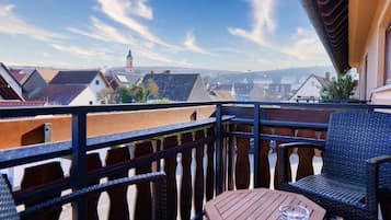 A balcony with wicker furniture and a table, overlooking a town with distinct rooftops and a church spire.