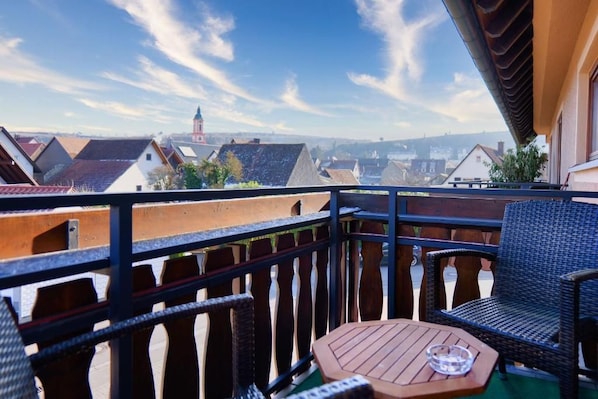 A balcony with wicker furniture and a table, overlooking a town with distinct rooftops and a church spire.
