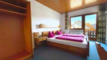 A bedroom with a bed, a nightstand, a wooden wardrobe, and a window with a view of buildings.