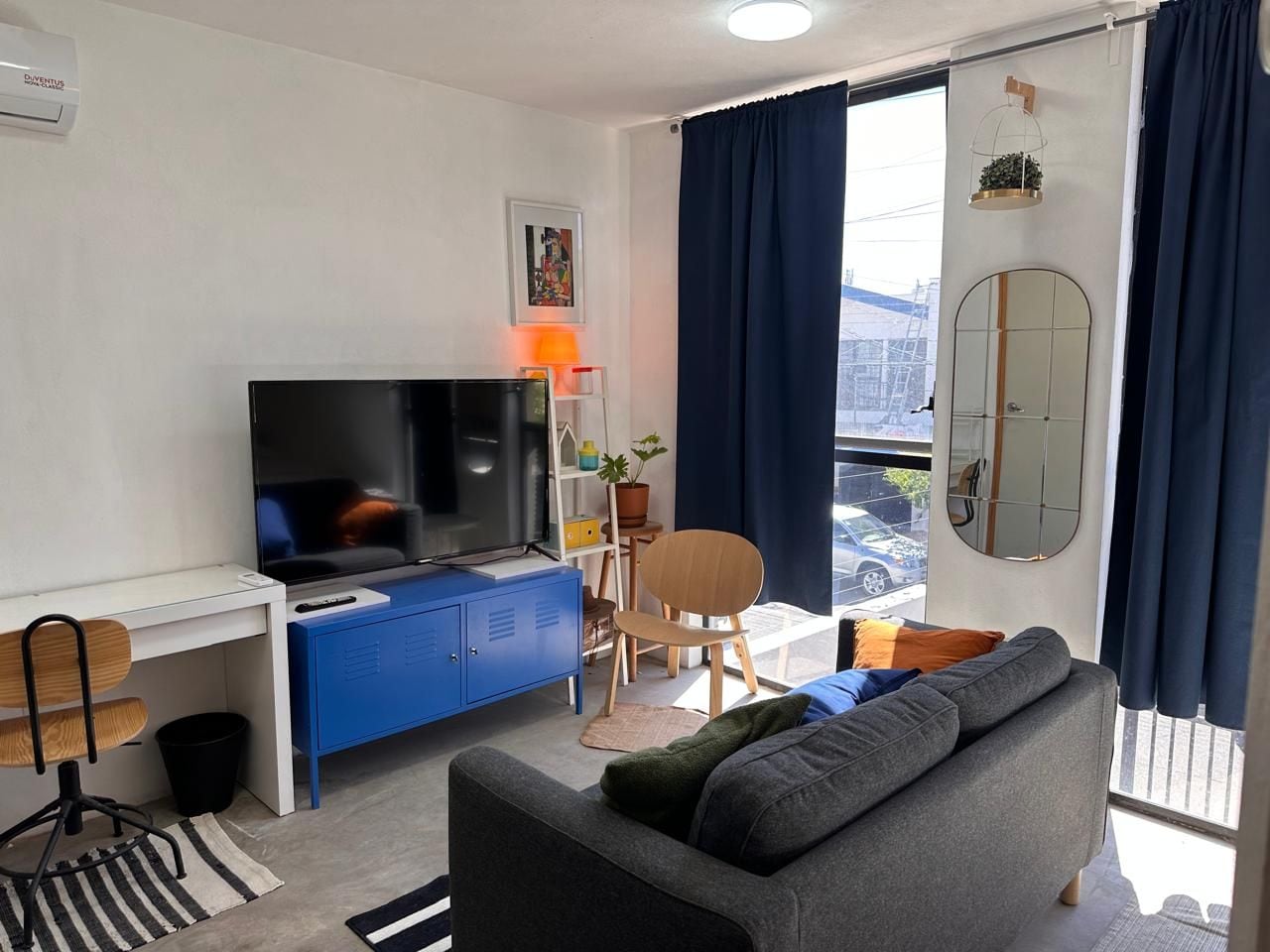 A modern living room with a flat-screen TV, a blue cabinet, a grey sofa, a dining table with chairs, and a balcony with a view.