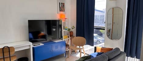 A modern living room with a flat-screen TV, a blue cabinet, a grey sofa, a dining table with chairs, and a balcony with a view.