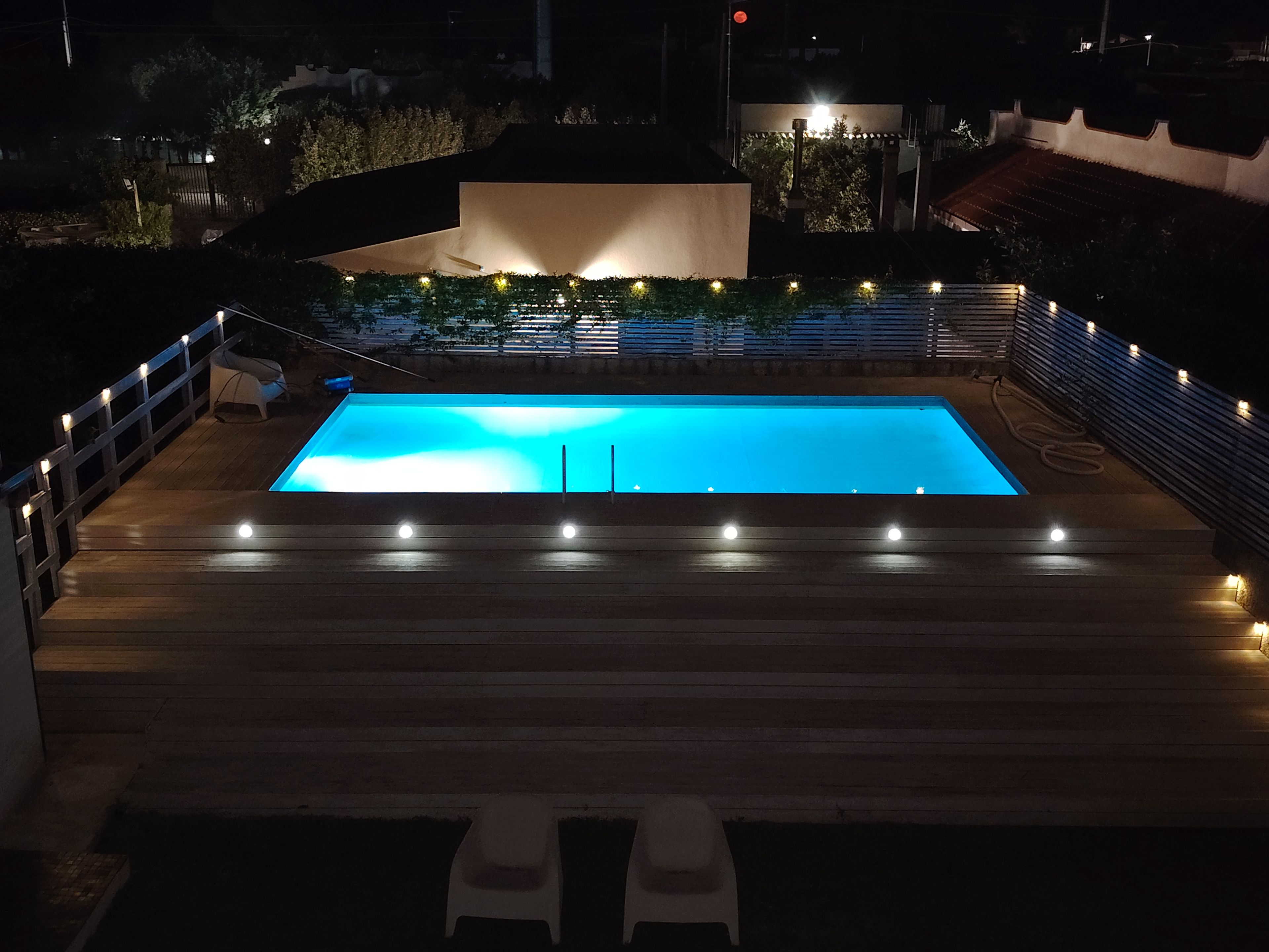 A rectangular swimming pool with a wooden deck surrounding it, equipped with lights and a few chairs.
