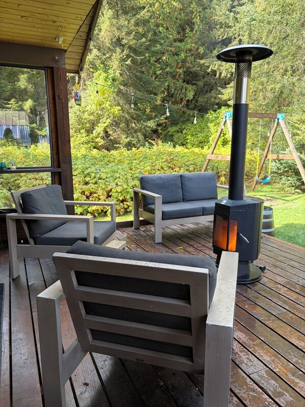 An outdoor seating area with a wood-burning stove, wooden deck, and a swing set in the background.
