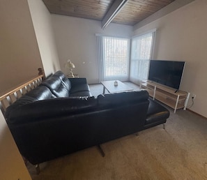 A living room with a black leather sofa, a coffee table, a flat-screen TV, and a window with blinds. - Lovely 2 bdrm Semi-detached (Calgary)