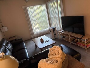 A living room with a black leather sofa, a coffee table with a remote control, a TV on a stand, and a bookshelf with books. - Lovely 2 bdrm Semi-detached (Calgary)