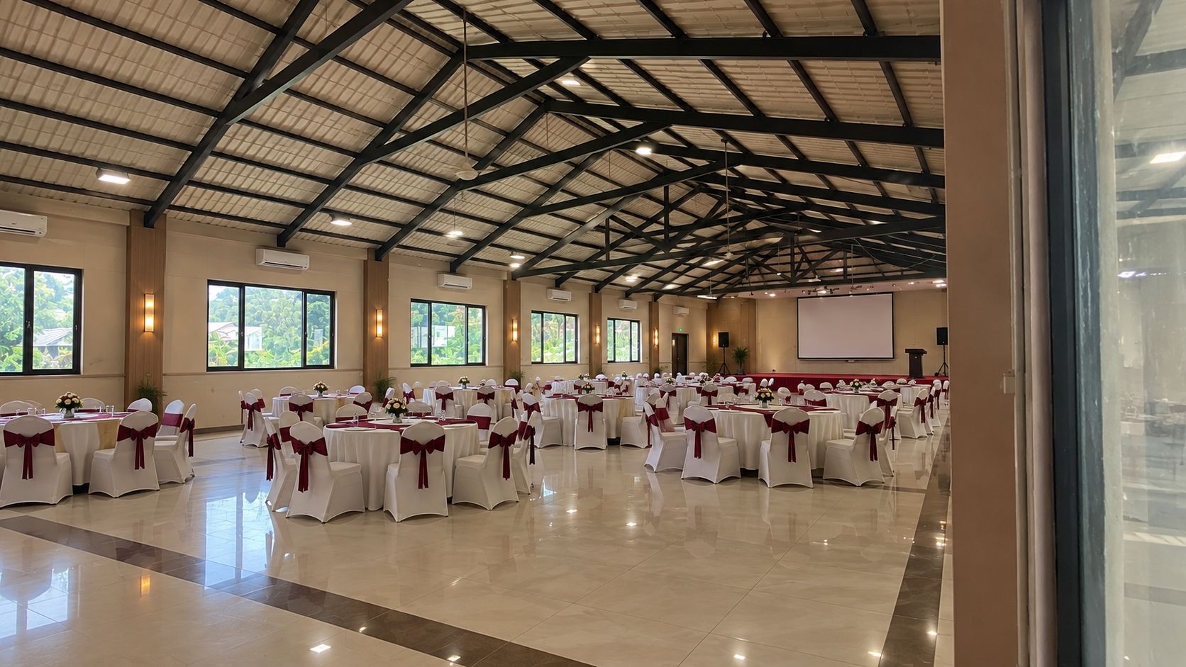 Un salón amplio con filas de mesas y sillas, unidades de aire acondicionado y un escenario con una pantalla.