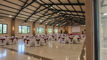 Un salón amplio con filas de mesas y sillas, unidades de aire acondicionado y un escenario con una pantalla.