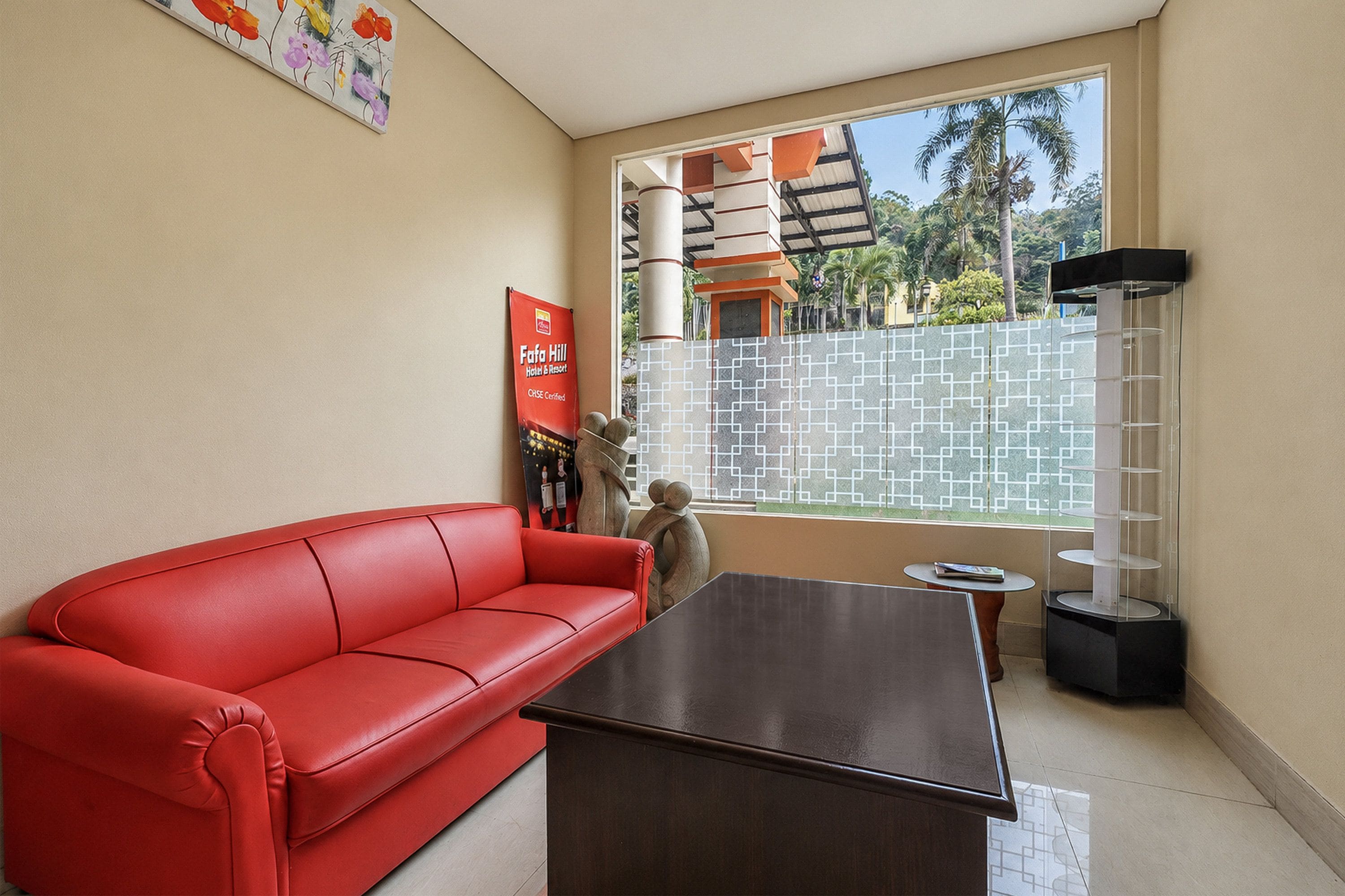 Un sofá de cuero rojo, una mesa de madera oscura y una escultura decorativa en una habitación con un ventanal grande, una obra de arte colgada en la pared y un aparador de vidrio.