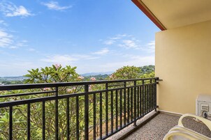 Un balcón con barandilla de metal, piso de baldosas y vistas a zonas verdes y montañas.