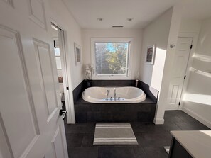 Un baño moderno con una bañera independiente, un ventanal con vista a árboles y piso cubierto de baldosas oscuras.
