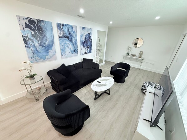 A modern living room with a black sofa, a black armchair, a white coffee table, and a flat-screen TV. There are abstract blue paintings on the wall and a small table with a plant.