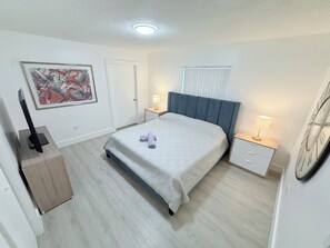 A bedroom with a bed, bedside tables, a TV, a painting, and a clock on the wall. - Modern 3BR Miami House, Sleeps 9, Gated Parking, Minutes to Wynwood (Miami)