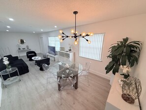 A modern living room with a glass dining table, black sofas, and a TV. - Modern 3BR Miami House, Sleeps 9, Gated Parking, Minutes to Wynwood (Miami)
