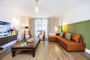 A modern living room with a wooden coffee table, two armchairs, a sofa, a television, and a large window with curtains. - Little Venice 2BR Retreat | Paddington & Hyde Park (London)