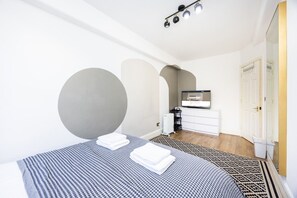 A bedroom with a bed, a TV, a white dresser, and a radiator. - Little Venice 2BR Retreat | Paddington & Hyde Park (London)