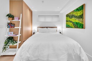 Une chambre à coucher avec un lit, un mur blanc, une armoire en bois, une étagère blanche avec des livres et une plante, une fenêtre avec des stores, et une œuvre d’art aux couleurs verte et jaune accrochée au mur.