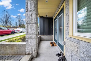 Une entrée d’immeuble avec un chemin d’accès en béton, une porte noire et une fenêtre avec des stores.