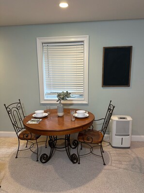 A dining area with a round table, four chairs, a window with blinds, a wall-mounted picture frame, and a standing heater. - Private Summerville Suite, Hot Tub, Gated 6 Acres, Near Charleston (Summerville)