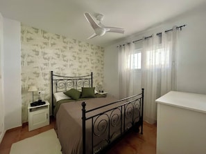Une chambre à coucher comprenant un lit, une table de chevet, un bureau, un ventilateur de plafond et une fenêtre avec des rideaux.