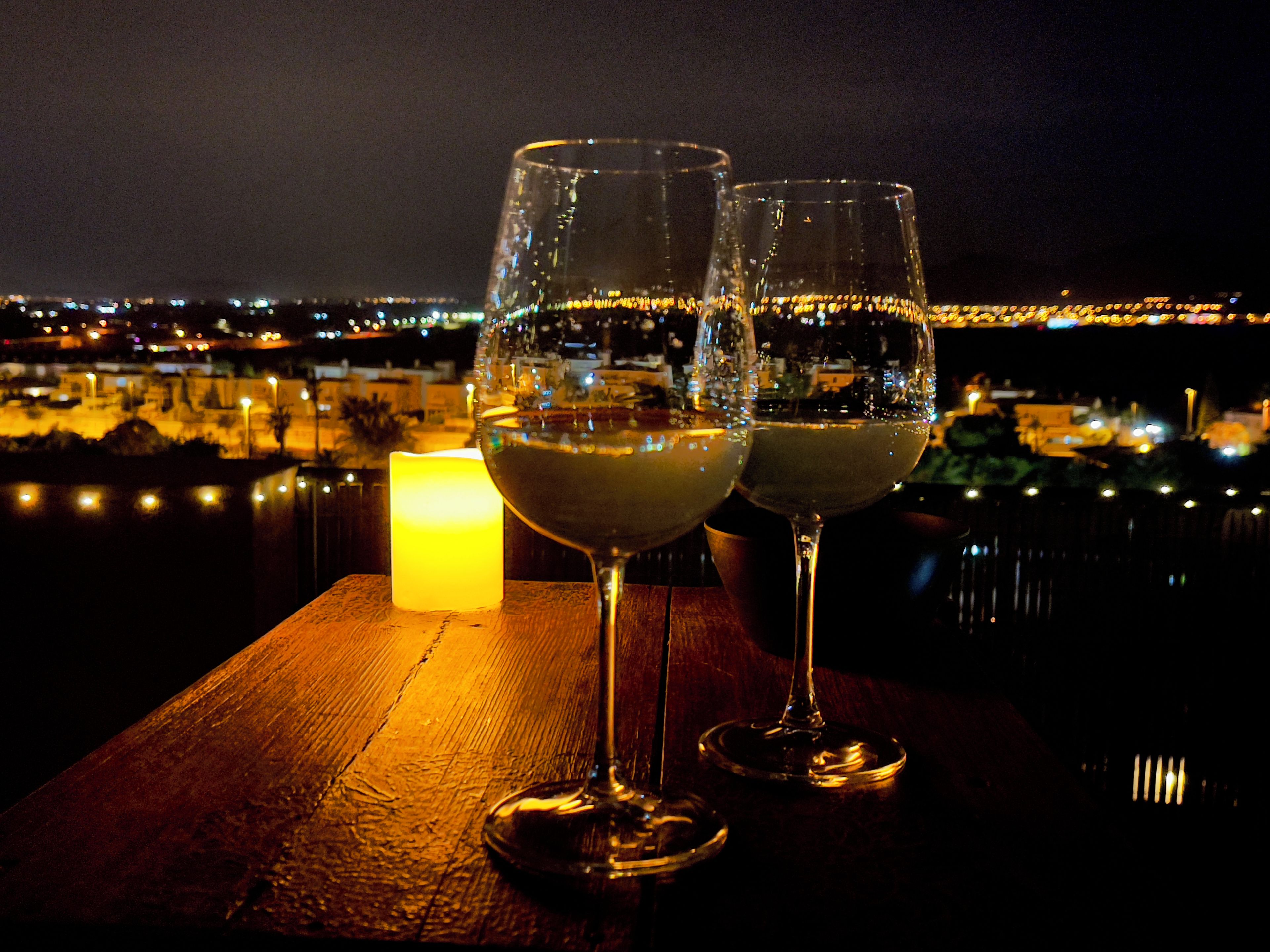 Deux verres de vin blanc sur une table en bois, avec une bougie allumée, donnant sur une vue nocturne de la ville.