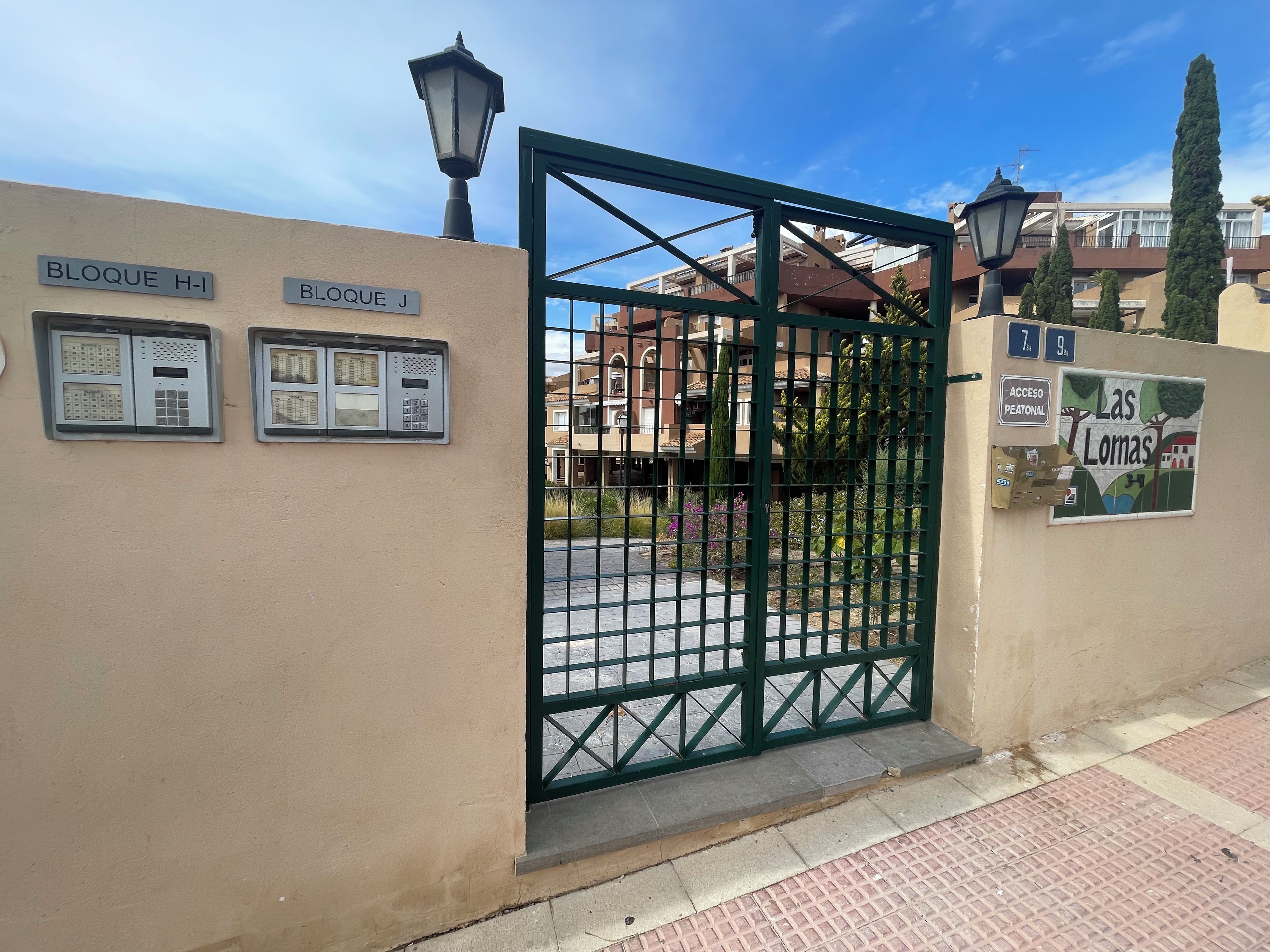A gated entrance with a metal gate, a street lamp, and a sign indicating 'BLOQUE H-1' and 'BLOQUE J'.