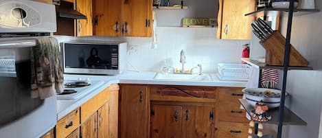 A kitchen with wooden cabinets, a microwave, a stove, a sink, and a shelf with kitchen items.