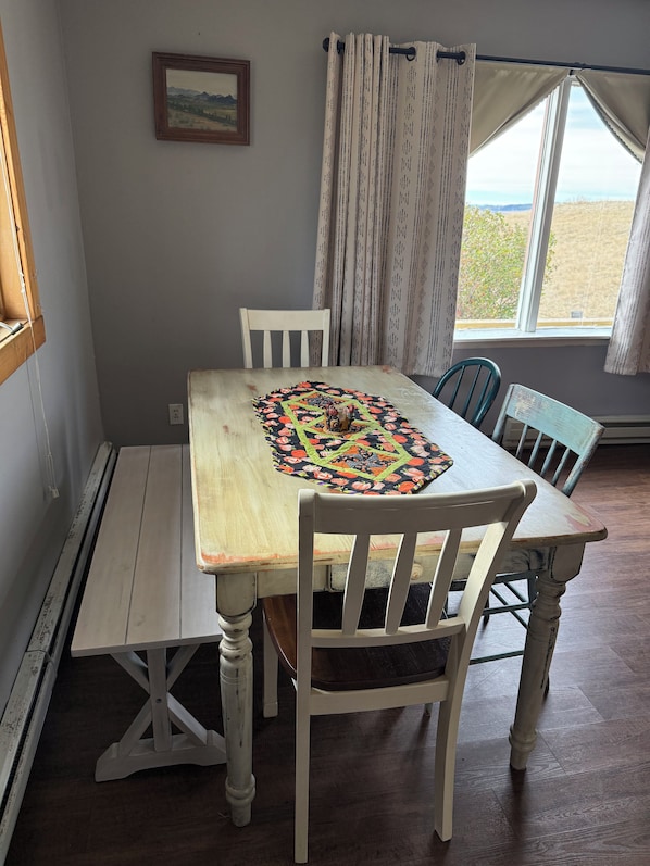 A dining area with a table, chairs, a bench, a window with curtains, and a wall-mounted picture.