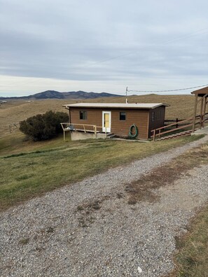 Una pequeña cabaña de madera con un camino de grava, una valla de madera y un campo de césped con colinas al fondo.