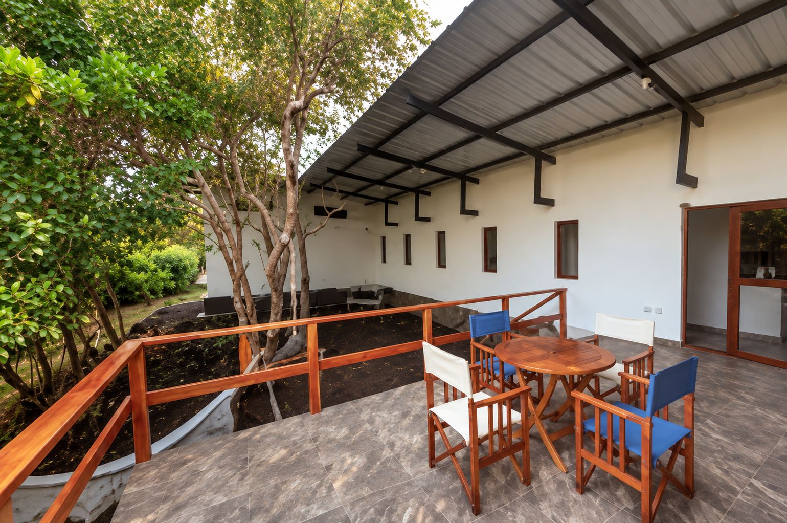 An outdoor patio with a wooden table and chairs, surrounded by a metal railing and a covered area with a sloped roof.