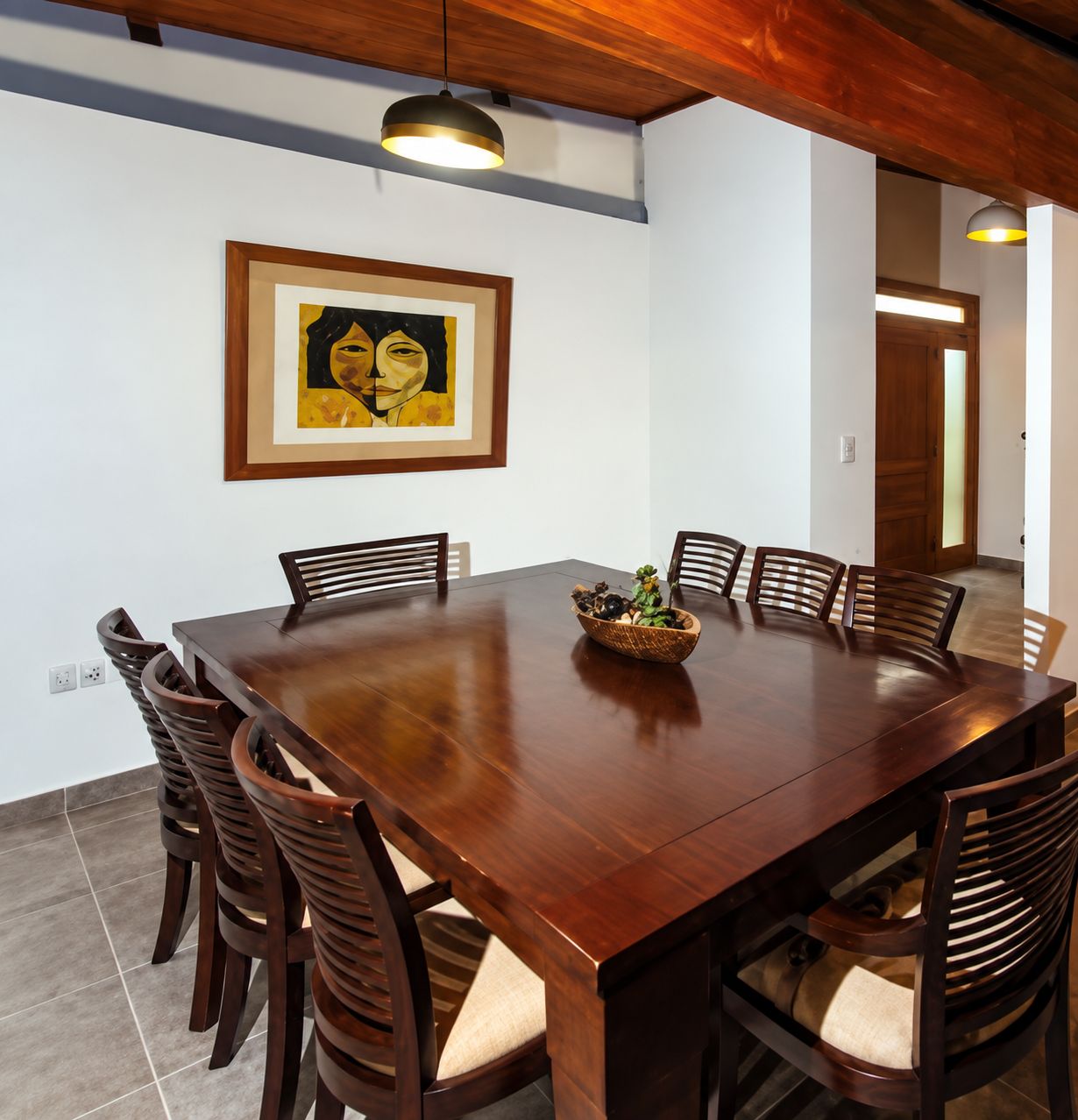 Une salle à manger avec une table et des chaises en bois, une œuvre d’art encadrée au mur et un panier de fruits sur la table.