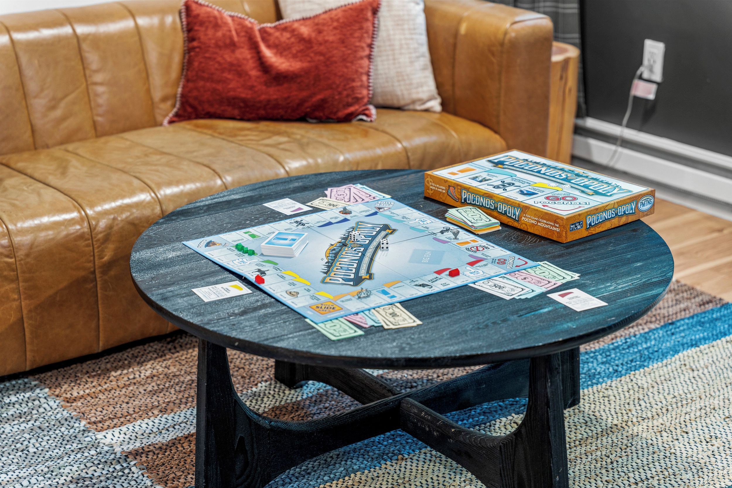 A board game titled 'Pocono's Opoly' on a round coffee table with a checkered rug and a brown leather couch in the background.