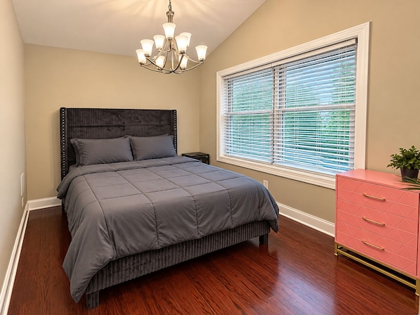 Une chambre à coucher comprenant un lit, une table de chevet, une commode, une fenêtre avec des stores et un lustre.