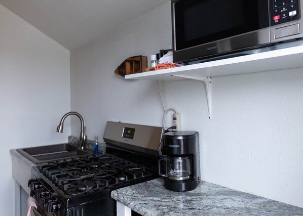 A compact kitchen with a gas stove, a coffee maker, a microwave, and a small shelf with kitchen items.