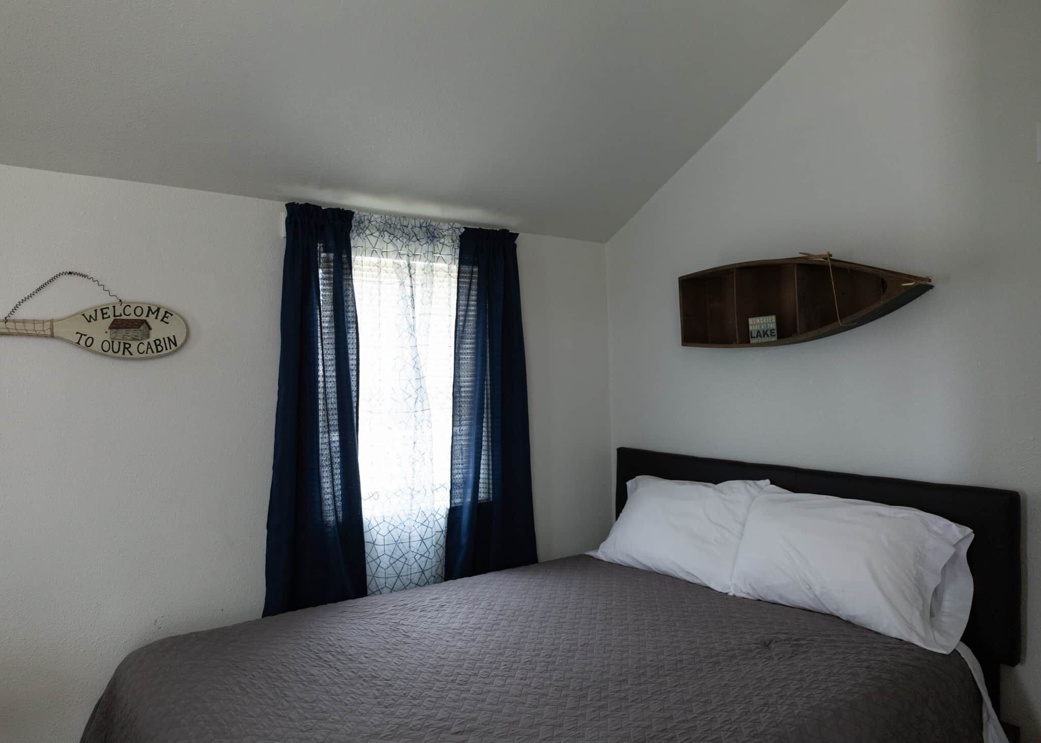 A bedroom with a bed, a window with curtains, a welcome sign, and a small boat decoration.
