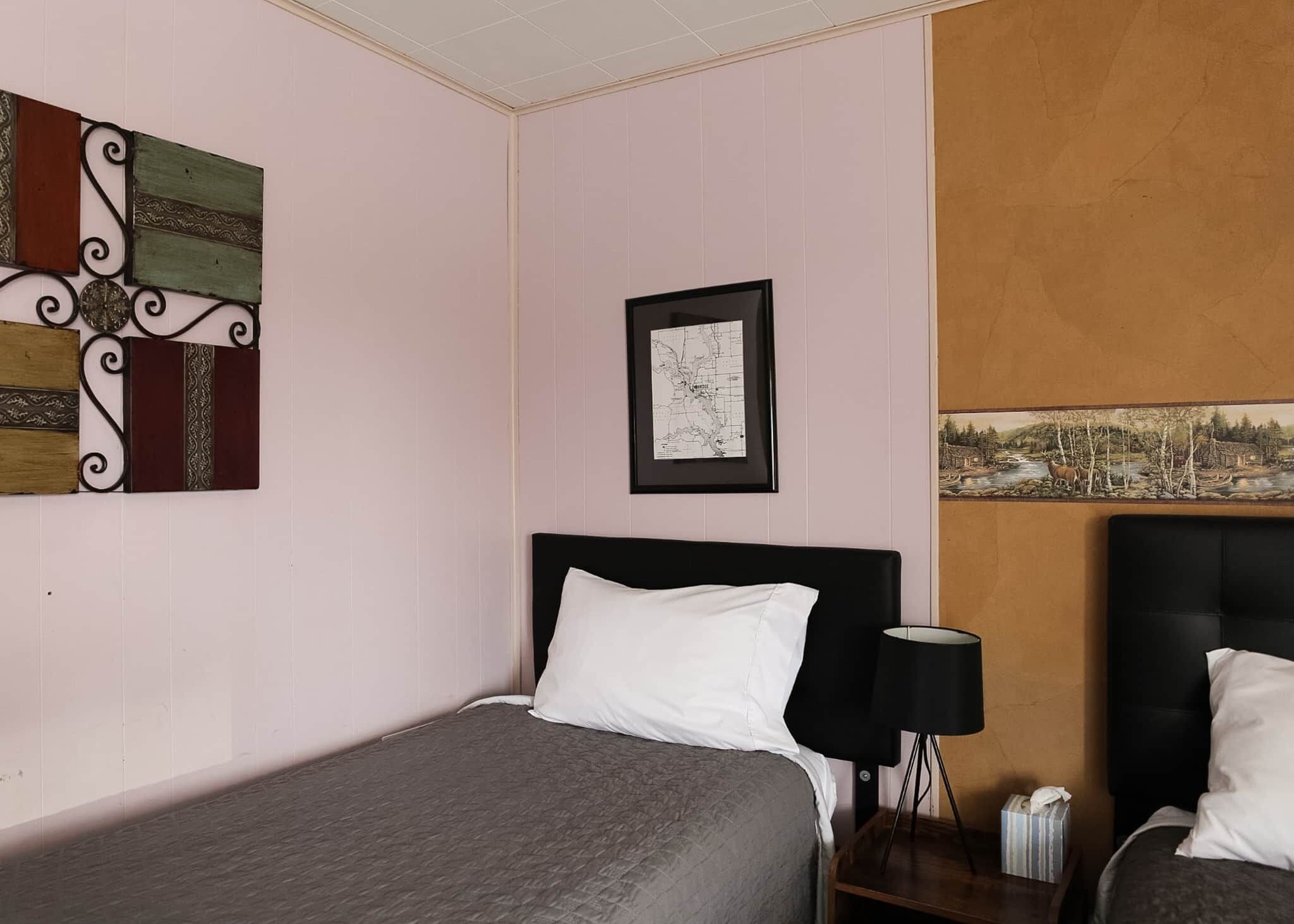 Hotellihuoneessa on sänky, yöpöytä, lamppu ja kaksi kehystettyä kuvaa seinällä.
