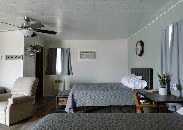 A hotel room with two beds, a chair, a desk, a ceiling fan, and an air conditioning unit.