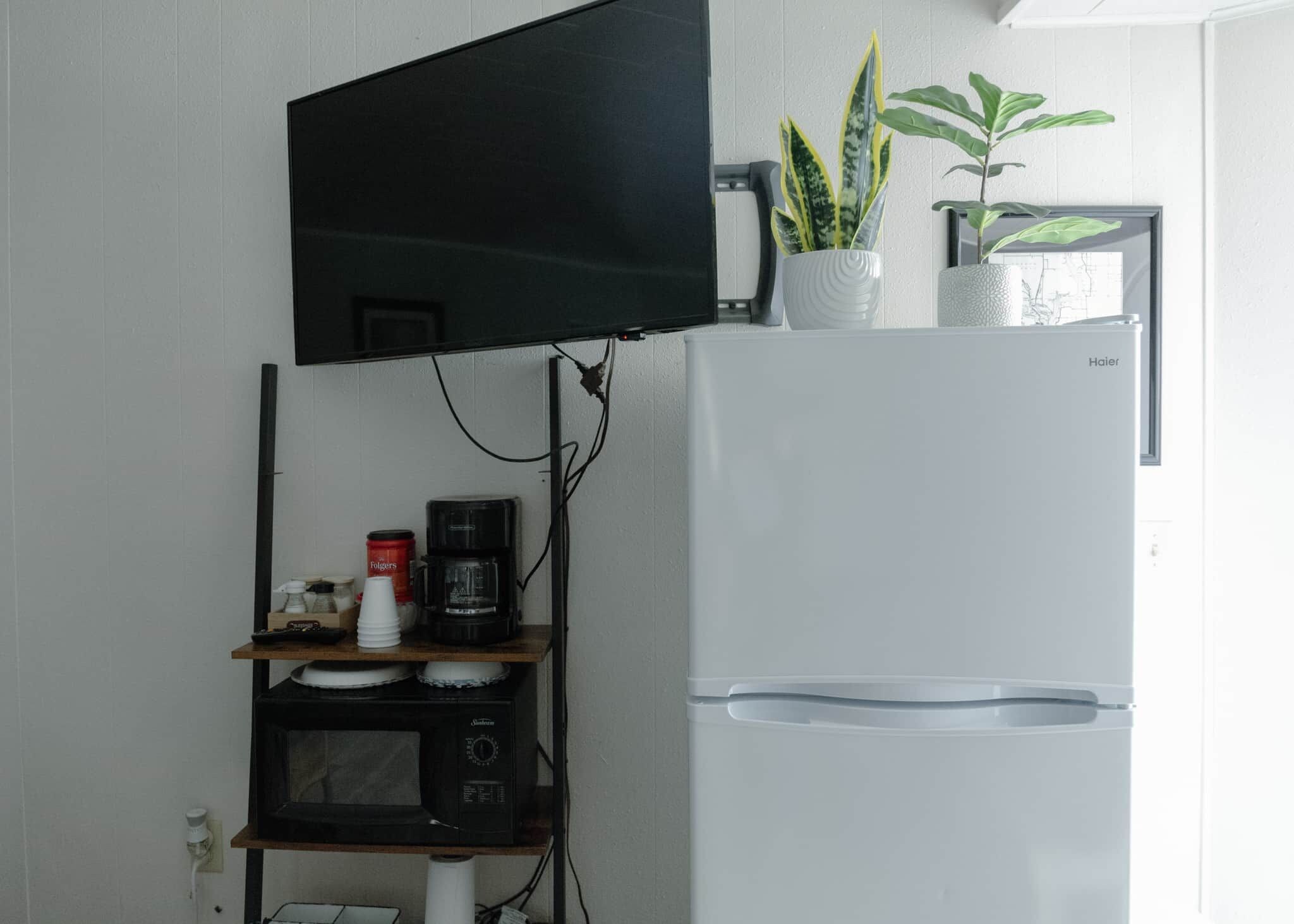 A kitchen with a flat-screen TV mounted above a microwave, a coffee maker, and a refrigerator with a water dispenser.